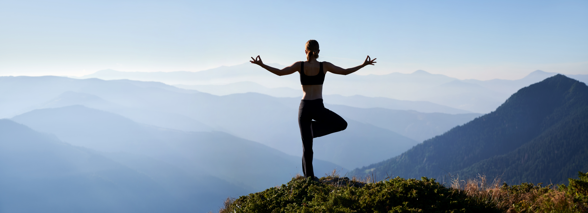 Frau macht stehende, einbeinige Yoga Pose auf einem Berg mit Panorama Aussicht